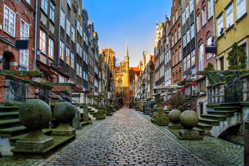 A cobblestone street in Gdańsk, Poland, lined with historic buildings adorned with ornate facades and amber shops, leads to the towering St. Mary's Church in the background. The street, known as Mariacka St, is famous for its charming architecture and picturesque atmosphere.
