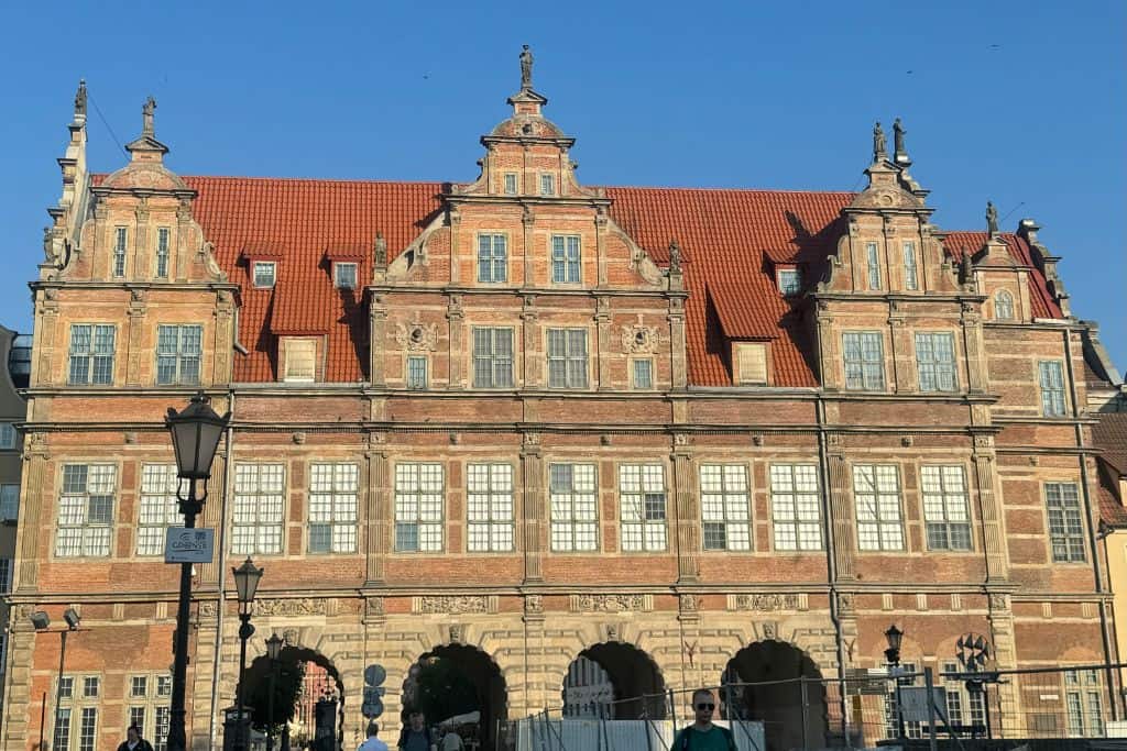 The Green Gate in Gdansk, a historic building with a red-tiled roof and ornate architectural details, stands prominently against a clear blue sky. People walk in front of the building, which features multiple windows and arched entrances at the ground level.