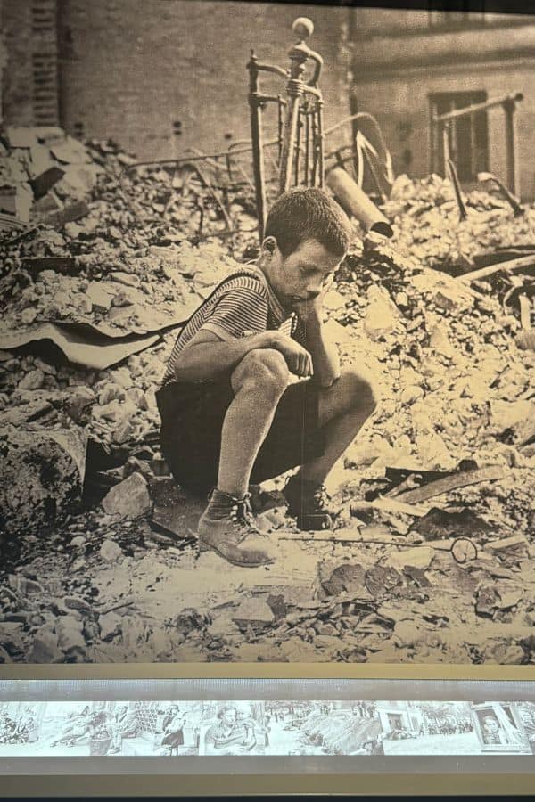 Black-and-white photo of a young boy sitting amidst rubble in a war-torn city, displayed at the Museum of the Second World War in Gdansk, Poland. The image reflects the devastation and human suffering caused by World War II.