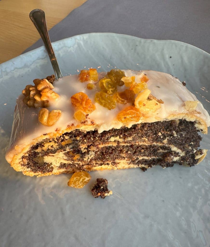 A slice of Polish dessert, makowiec, is shown on a light blue plate. The dessert is a poppy seed roll topped with white icing, walnuts, and dried fruits such as raisins and candied orange peel.