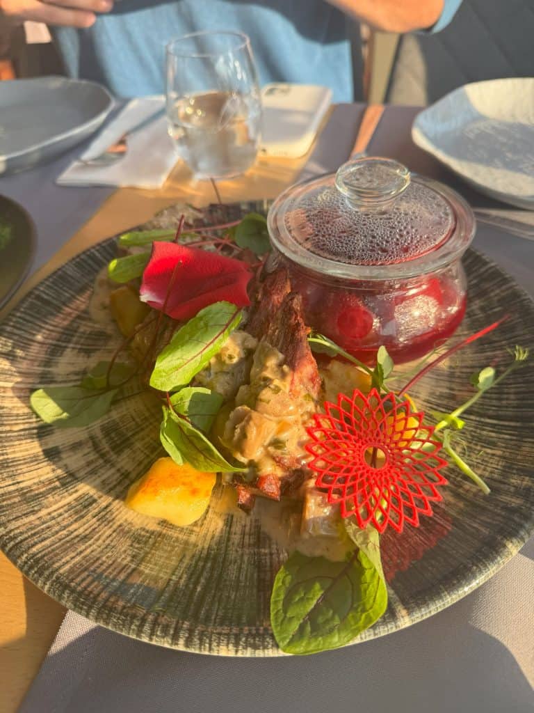 A plate featuring wild boar sausage, pierogi, fresh greens, and a red decorative piece, accompanied by a glass jar of pickled beetroot.