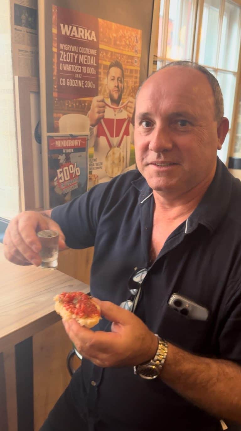 A person is holding a shot glass filled with vodka, in one hand and a piece of bread topped with steak tartare in the other hand.