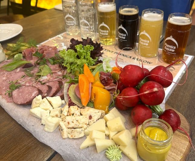 A Polish charcuterie board featuring an assortment of sliced meats, various cheeses, fresh vegetables including radishes and bell peppers, leafy greens, and a small jar of mustard. In the background, there are four glasses of different types of beer.