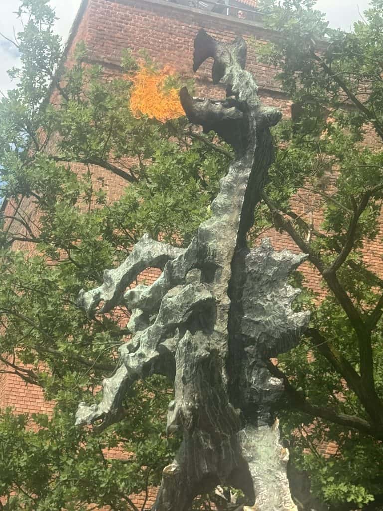 Dragon statue near Wawel Castle in Krakow, breathing fire, with a brick building and trees in the background.