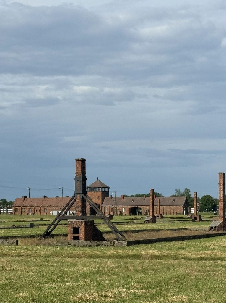 Ruins of brick chimneys and barracks at Birkenau concentration camp, with the main entrance gate visible in the distance.